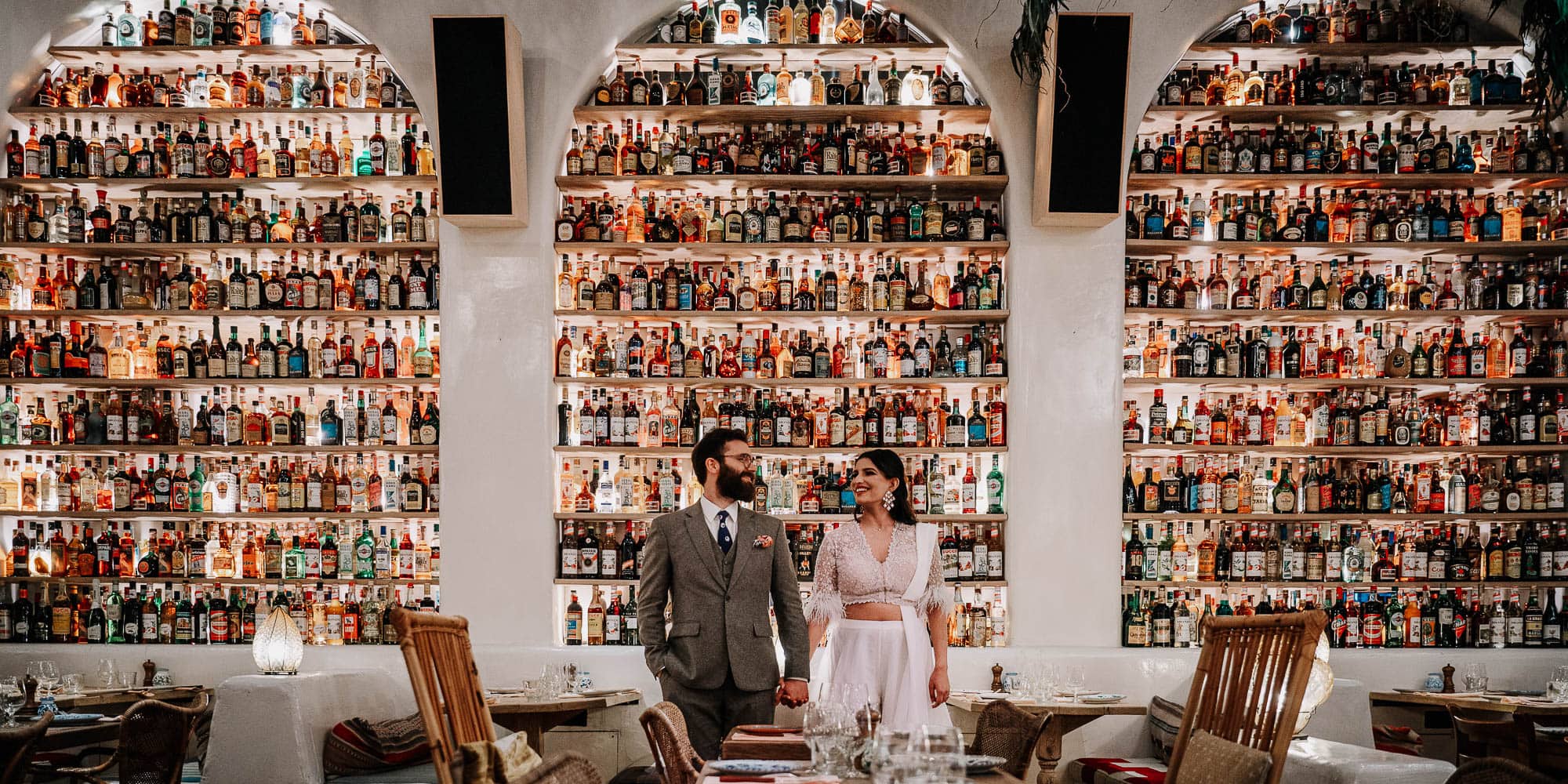 A Couple stands in front of lots of bottle of alcohol in a restaurant, Holding hands