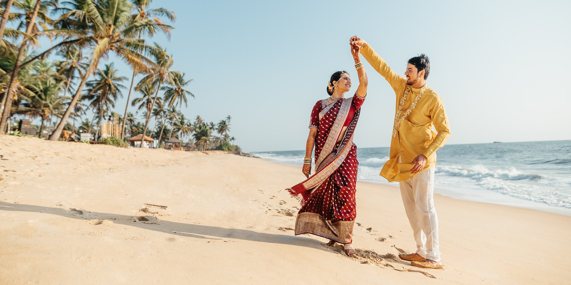 A South Indian couple walking and dancing on the beaching in Mangalore India. The groom is wearing yellow and the bride is wearing a red sari